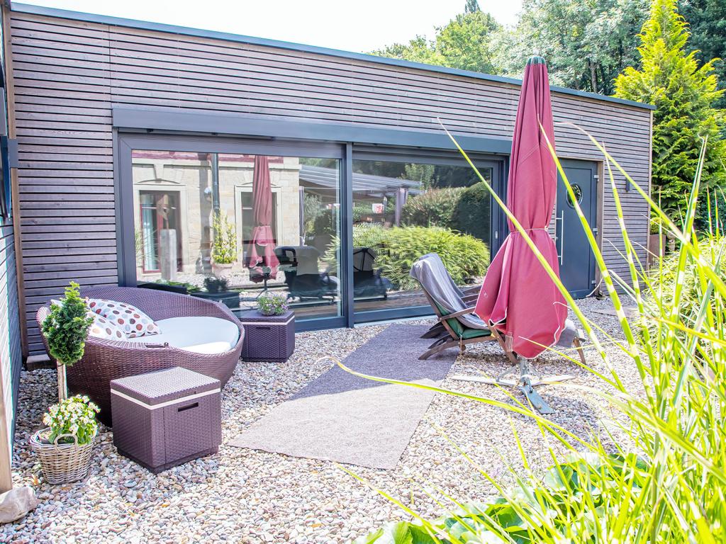 Moderner Garten mit Kiesboden, Lounge-Möbeln aus Rattan, zwei geschlossenen Sonnenschirmen und großem Fensterfront eines Gebäudes