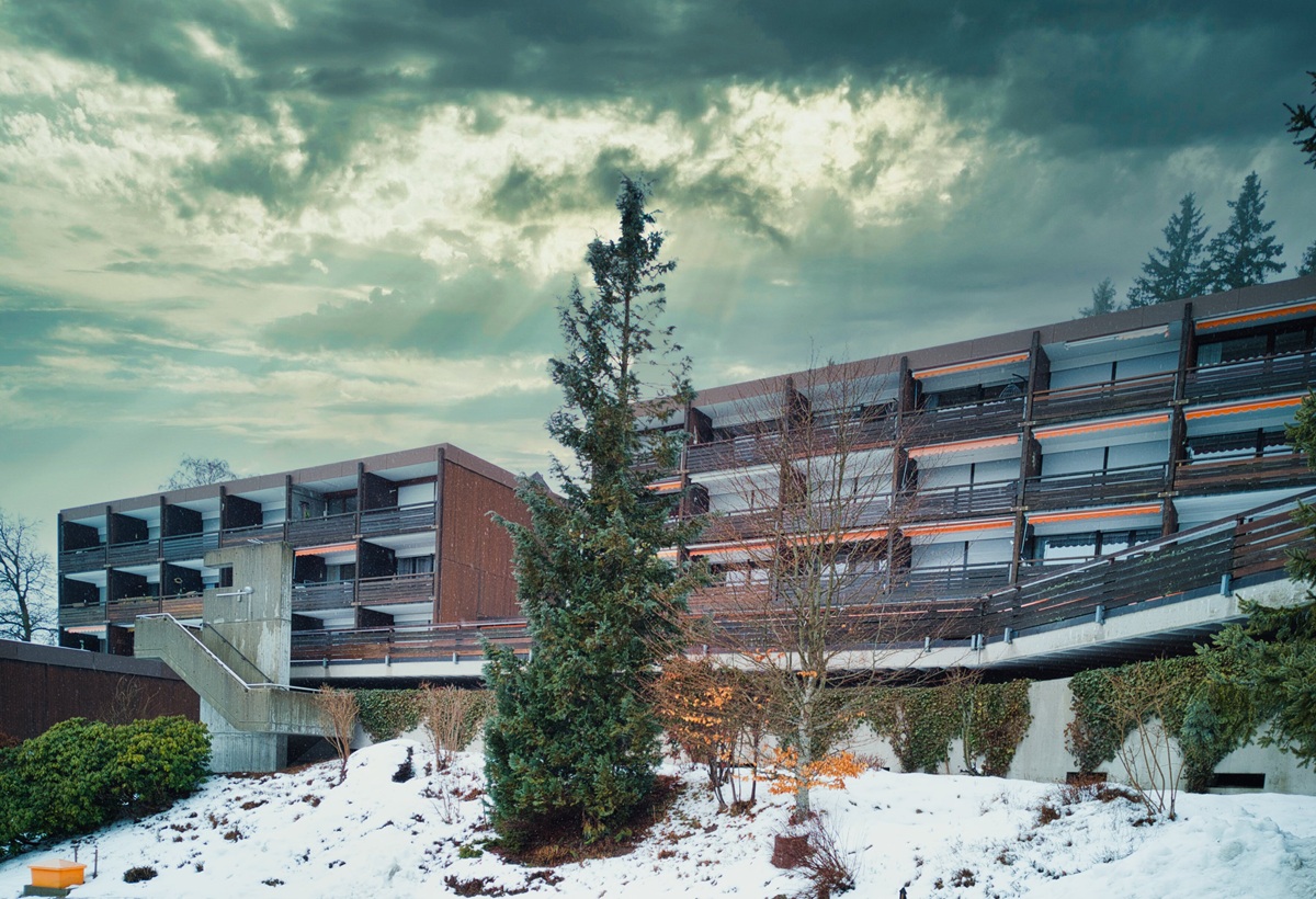 Mehrstöckiges Gebäude mit Holzfassade und Balkonen vor bewölktem Himmel und schneebedecktem Boden mit Nadelbäumen. Das sind Ferienappartements in Schonach, Schwarzwald. Präsentiert von Schwebius-Immobilien