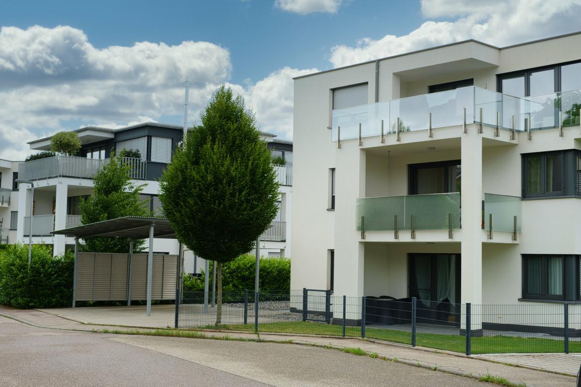 Schwebius-Immobilien bietet an: Mehrfamilienhaus mit Balkonen und Glasgeländern an einer ruhigen Straße mit Baum und Carport